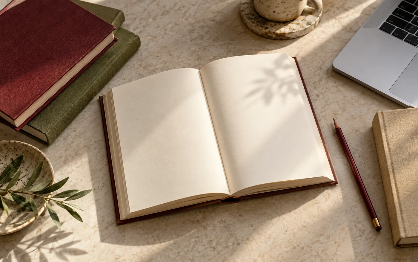 An open book, plain clothbound books, and a laptop on a sunlit table
